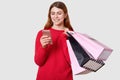 Young beautiful fashionable Caucasian woman holds shopping bags in one hand and smartphone in another isolated on white background
