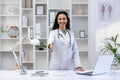 Young beautiful female doctor smiling and looking at camera standing inside clinic office, extending hand forward Royalty Free Stock Photo