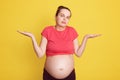 Young beautiful female expecting baby, stands isolated over yellow background, being clueless and confused with open arms, having