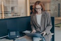 Beautiful redhead businesswoman sitting on top of desk and using laptop, working alone in office Royalty Free Stock Photo