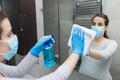 Young beautiful female wearing protective face mask and cleaning mirror in bathroom. Coronavirus protection prevention