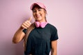 Young beautiful fitness sports woman wearing training cap and headphones over pink background doing happy thumbs up gesture with Royalty Free Stock Photo