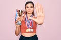 Young beautiful fitness winner athlete woman wearing sport medals and trophy with open hand doing stop sign with serious and