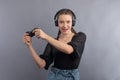 Young beautiful gamer girl playing video game using joystick and headphones on grey background. Girl in casual wear with emotional