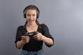 Young beautiful gamer woman playing video game using joystick and headphones on grey background. Woman in casual wear with