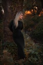 The young beautiful girl in a black dress stands in the wood near a big tree at a stream at sunset Royalty Free Stock Photo