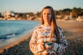 Young beautiful girl in cozy sweater holding coffee cup while enjoying winter sun on seaside shore during mild sunset Royalty Free Stock Photo