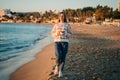 Young beautiful girl in cozy sweater holding coffee cup while enjoying winter sun on seaside shore during mild sunset Royalty Free Stock Photo