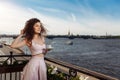 Young beautiful girl with curly hair in a pink dress is standing on the balcony and drinking coffee. Happy woman smiling and Royalty Free Stock Photo