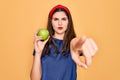 Young beautiful girl eating fresh organic healthy green apple over yellow background pointing with finger to the camera and to Royalty Free Stock Photo