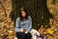 Young beautiful girl in a gray jacket sitting in the autumn forest near a large tree with a mobile phone Royalty Free Stock Photo