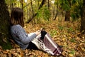 Young beautiful girl in a gray jacket sitting in the autumn forest near a large tree with a mobile phone Royalty Free Stock Photo