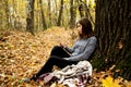 Young beautiful girl in a gray jacket sitting in the autumn forest near a large tree with a mobile phone Royalty Free Stock Photo