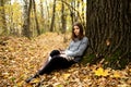 Young beautiful girl in a gray jacket sitting in the autumn forest near a large tree with a mobile phone Royalty Free Stock Photo