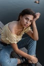 Young beautiful girl hipster model in a yellow blouse and blue jeans sits on the pier near the water Royalty Free Stock Photo