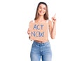 Young beautiful girl holding act now banner surprised with an idea or question pointing finger with happy face, number one