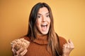 Young beautiful girl holding bowl of healthy cornflakes cereals over yellow background screaming proud and celebrating victory and