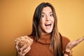 Young beautiful girl holding bowl of healthy cornflakes cereals over yellow background very happy and excited, winner expression