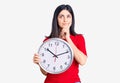 Young beautiful girl holding clock serious face thinking about question with hand on chin, thoughtful about confusing idea Royalty Free Stock Photo