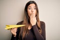 Young beautiful girl holding paper plane standing over isolated white background cover mouth with hand shocked with shame for Royalty Free Stock Photo