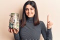 Young beautiful girl holding tips dollars jar smiling with an idea or question pointing finger with happy face, number one