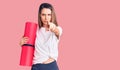 Young beautiful girl holding yoga mat pointing with finger to the camera and to you, confident gesture looking serious Royalty Free Stock Photo