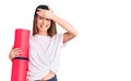 Young beautiful girl holding yoga mat stressed and frustrated with hand on head, surprised and angry face Royalty Free Stock Photo