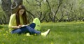 Young beautiful girl with long dark hair sitting in nature on a green field with dandelions under a tree, reading a book Royalty Free Stock Photo