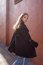 Young beautiful girl with long hair in black coat on sunny day turns around against the red wall. Street style portrait Royalty Free Stock Photo