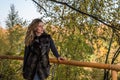 Young beautiful girl with long hair, in a fur jacket, stands on a wooden railing on a bright sunny day while walking in the autumn Royalty Free Stock Photo