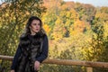 Young beautiful girl with long hair, in a fur jacket, stands on a wooden railing on a bright sunny day while walking in the autumn Royalty Free Stock Photo