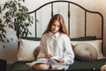 A young beautiful girl with long hair in a white shirt sits on the bed and reads a book Royalty Free Stock Photo