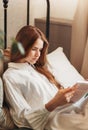 A young beautiful girl with long hair in a white shirt sits on the sofa and reads a book. Vertical photo Royalty Free Stock Photo