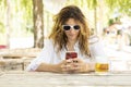 Young beautiful girl in the middle of summer sitting at a table with a beer in nature while looking at her phone smiling Royalty Free Stock Photo