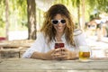 Young beautiful girl in the middle of summer sitting at a table with a beer in nature while looking at her phone smiling Royalty Free Stock Photo