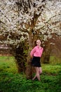 Young beautiful girl in a pink shirt standing under blossoming apple tree and enjoying a sunny day.