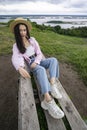 A young and beautiful girl sits on a wooden bench, on a high bank overlooking a large river