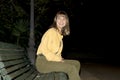 Young beautiful girl sitting in a bench in a public city park at night while looking to camera