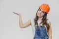 A young beautiful girl stands on a gray background. It is dressed in an orange helmet on the head. During this, he has a Royalty Free Stock Photo