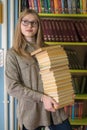 Young beautiful girl student holds in her hands a large stack of books in the library against the background of bookshelves Royalty Free Stock Photo