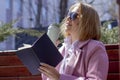 A young beautiful girl in sunglasses drinks coffee from an eco-friendly thermocup and holds a notebook while sitting on a bench Royalty Free Stock Photo