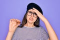 Young beautiful girl wearing glasses and fashion beret holding french sweet pastry macaron stressed with hand on head, shocked