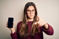 Young beautiful girl wearing glasses showing smartphone over isolated white background pointing with finger to the camera and to Royalty Free Stock Photo