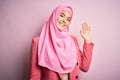 Young beautiful girl wearing muslim hijab standing over isolated pink background Waiving saying hello happy and smiling, friendly