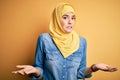 Young beautiful girl wearing muslim hijab standing over isolated yellow background clueless and confused expression with arms and Royalty Free Stock Photo