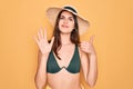 Young beautiful girl wearing swimwear bikini and summer sun hat over yellow background showing and pointing up with fingers number Royalty Free Stock Photo