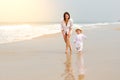 A young beautiful girl in a white shirt runs along a sandy beach in the sea with her son in white clothes and hat