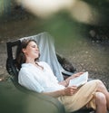 Young beautiful girl woman enjoys nature and reading a book in the summer in the garden of her house sitting on a chair Royalty Free Stock Photo