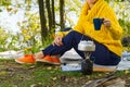 Young beautiful girl in a yellow sweater makes coffee in the forest on a gas burner. Making coffee on a primus stove in the autumn