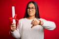 Young beautiful graduate asian woman holding university degree diploma over red background with angry face, negative sign showing Royalty Free Stock Photo